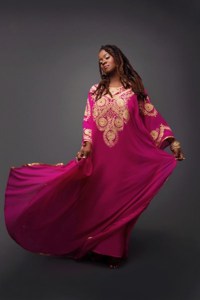 Black lady barefoot dancing in a flowing bright pink and elaborately gold decorated dress against a grey background