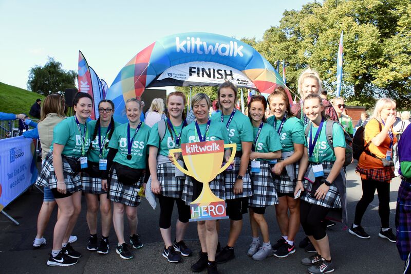 Group of women wearing kilts and bliss t-shirts at Kiltwalk finish line holding foamboard cut-out Kiltwalk hero trophy