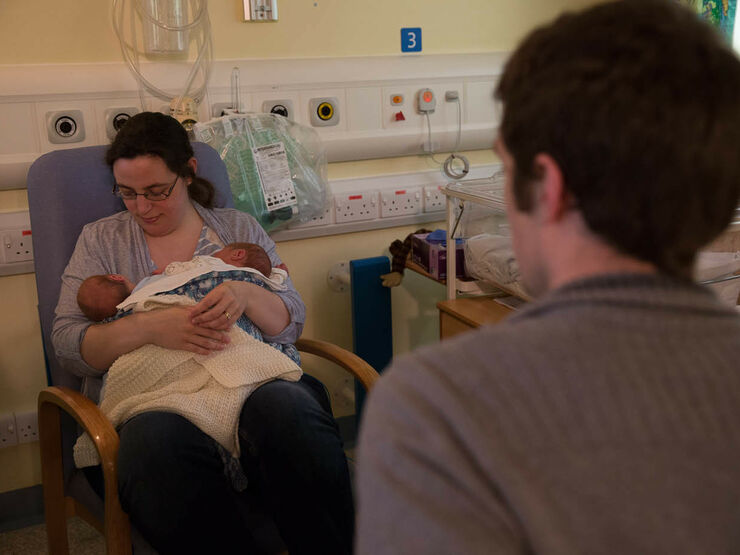 Dad in foreground looking away from camera towards Mum who is sitting holding and looking at her twins in her arms