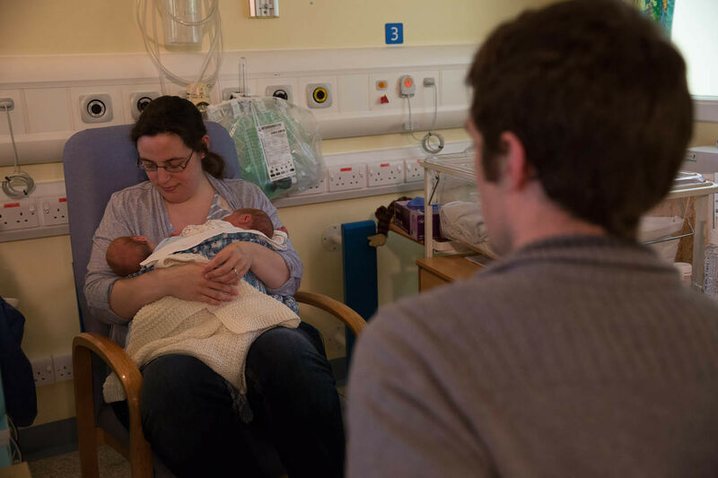 Dad in foreground looking away from camera towards Mum who is sitting holding and looking at her twins in her arms