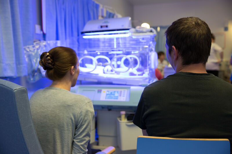 Mum and Dad on the neonatal unit sat down with their backs to the camera, looking towards an incubator and blue light from phototherapy treatment.