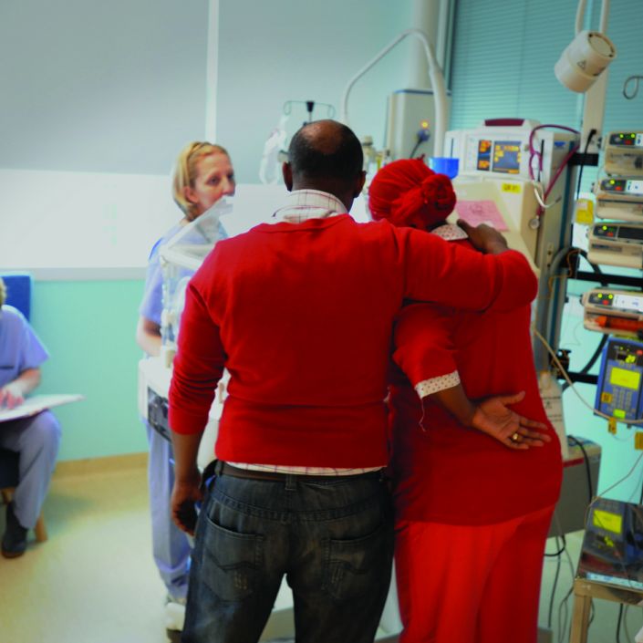 Facing away from the camera, Dad puts arm around Mum in a caring way whilst looking at baby in an incubator surrounded by two health professionals