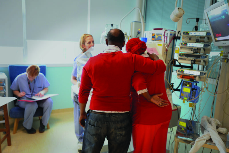 Facing away from the camera, Dad puts arm around Mum in a caring way whilst looking at baby in an incubator surrounded by two health professionals
