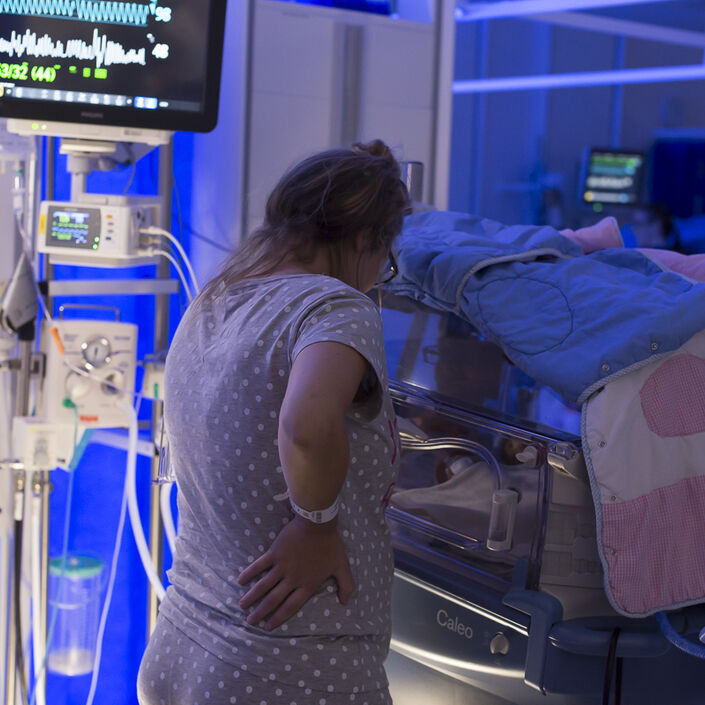 Mum standing by incubator in low light, looking at her baby and away from the camera