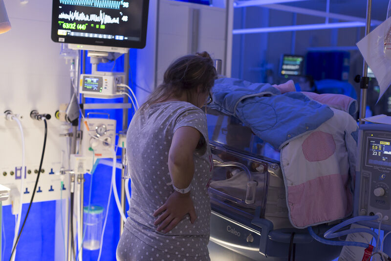 Mum standing by incubator in low light, looking at her baby and away from the camera