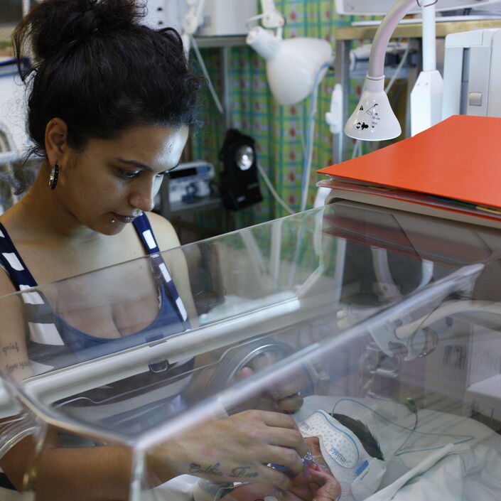 Mum standing looking into incubator at her baby who is wearing an eye mask. She is reaching through the incubator to comfort her baby.
