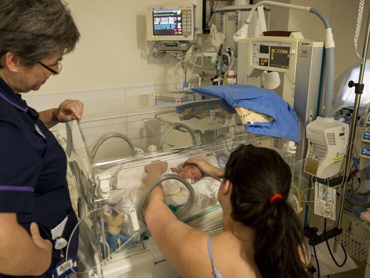 Mum sitting down looking away from the camera reaching into the incubator with both hands, holding her baby and looking at eye level. A nurse is standing next to her looking into the incubator