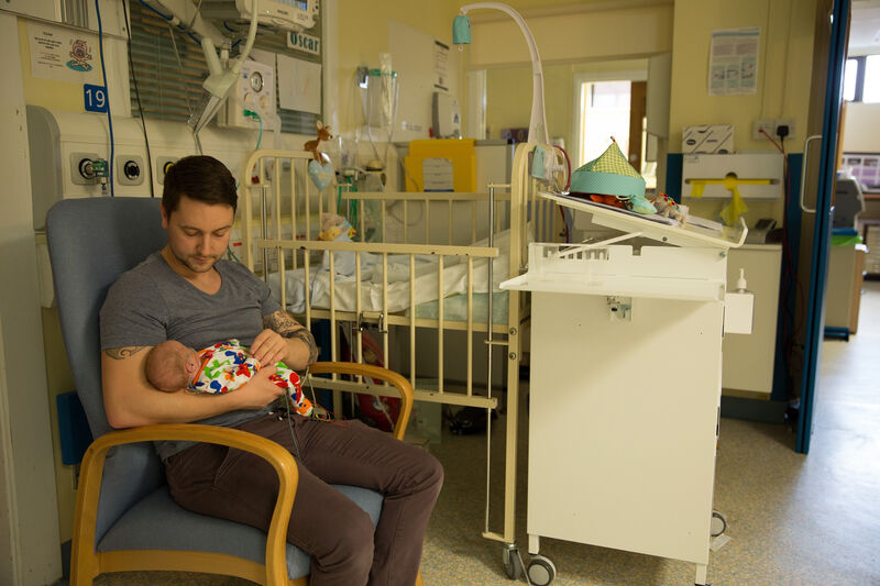 Father holding his baby in a chair next to a cot in an neonatal unit
