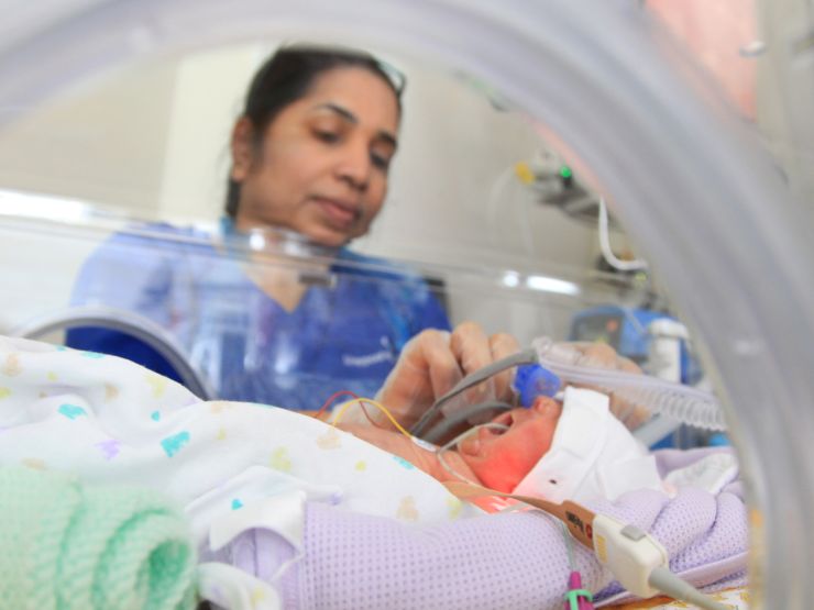 Baby in incubator on neonatal unit with nurse in background attending to the baby
