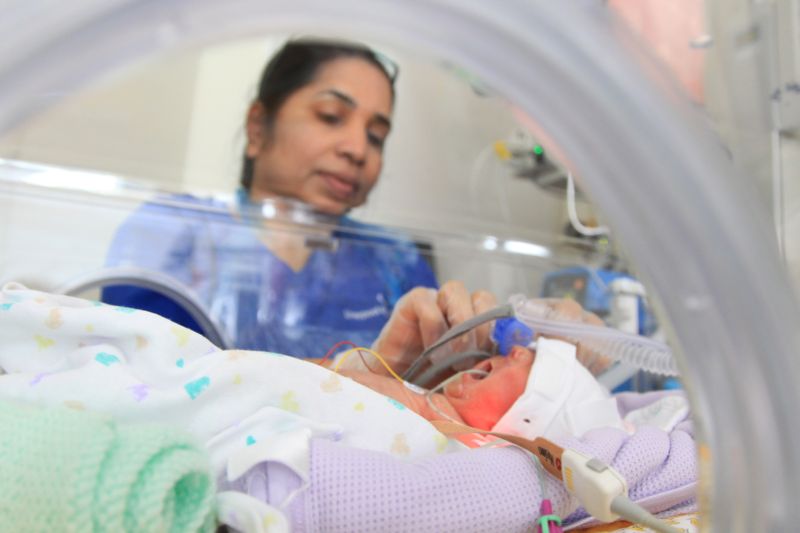 Baby in incubator on neonatal unit with nurse in background attending to the baby