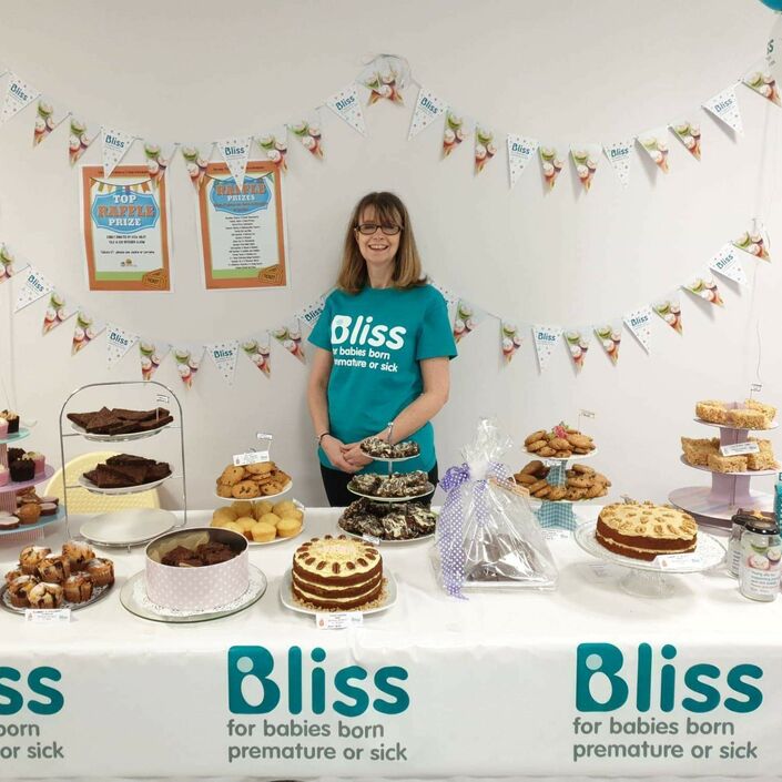 Lady wearing Bliss t-shirt standing behind a table of cakes and bunting and balloons behind, and table decorated with Bliss logo