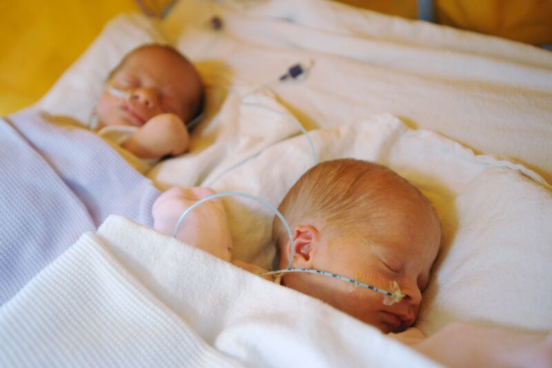 Two babies laying side by side wrapped in blankets and sleeping, with tubes attached to their nose
