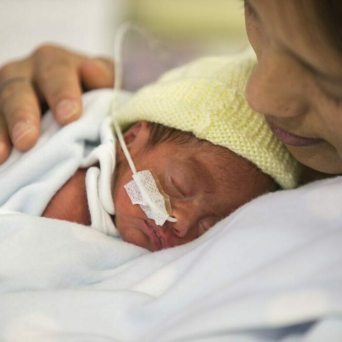 A mother leaning back with her baby sleeping on her chest. Baby is wearing a knitted, yellow bonnet and has a nasogastric tube.