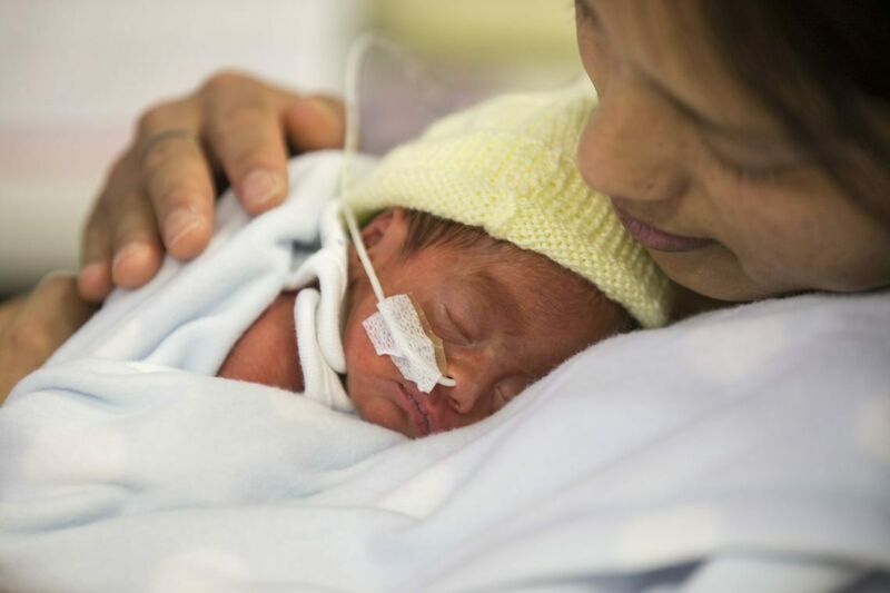 A mother leaning back with her baby sleeping on her chest. Baby is wearing a knitted, yellow bonnet and has a nasogastric tube.