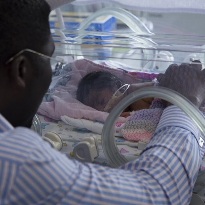 Dad with hand on baby in incubator with baby lying on front asleep and blanket over