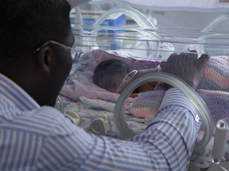 Dad with hand on baby in incubator with baby lying on front asleep and blanket over