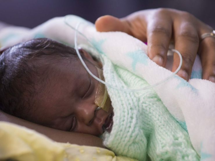 A baby sleeps on his mother's chest, wrapped in blankets, with a nasogatric tube taped to his face
