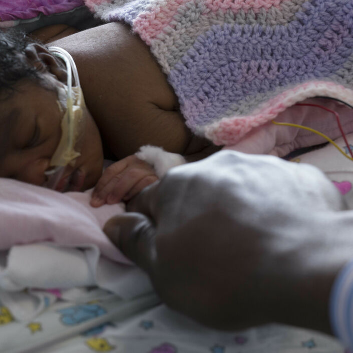 Baby with tubes in nose covered in a blanket holding their father's finger