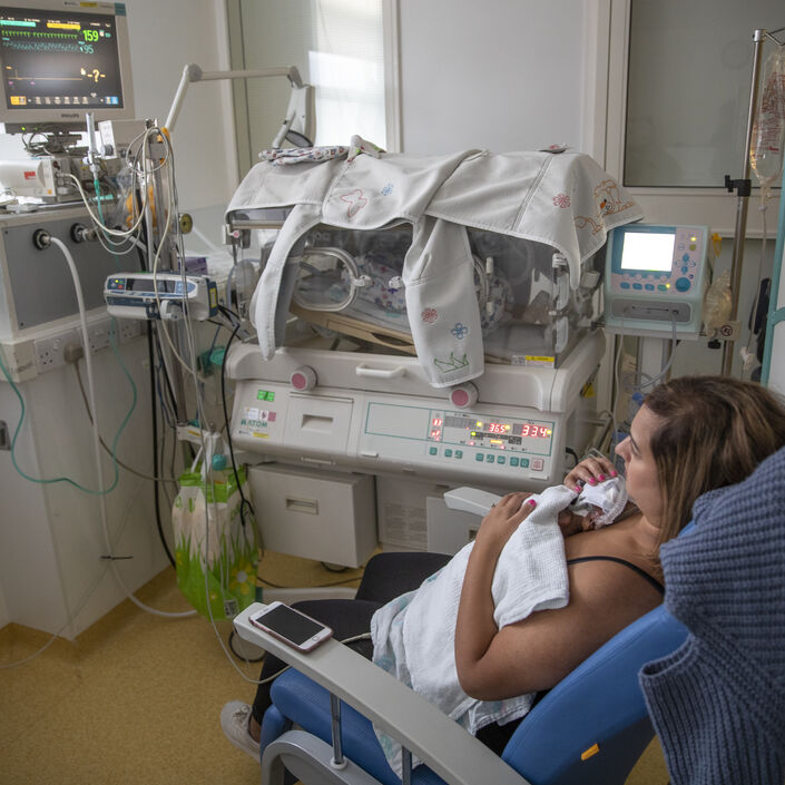 Mum sat on a hospital chair with her baby resting on her chest. In the background is an incubator and monitor