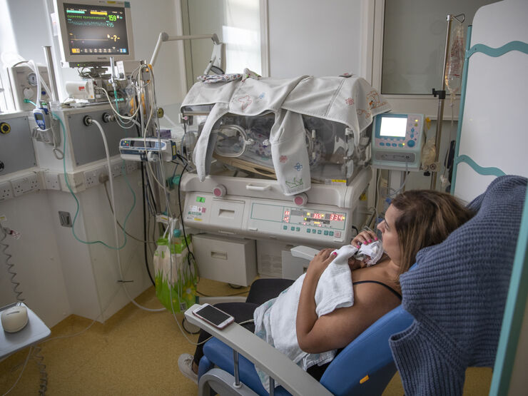 Mum sat on a hospital chair with her baby resting on her chest. In the background is an incubator and monitor