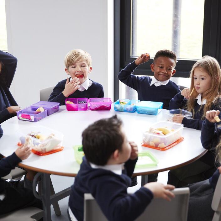 Group of primary school children sat at a round table having lunch and laughing