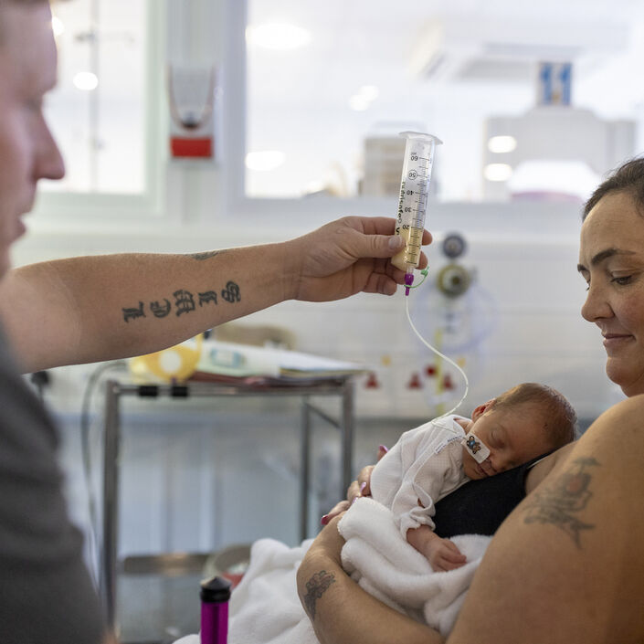 Mum holding baby on her chest while dad holds feeding tube in the air above baby