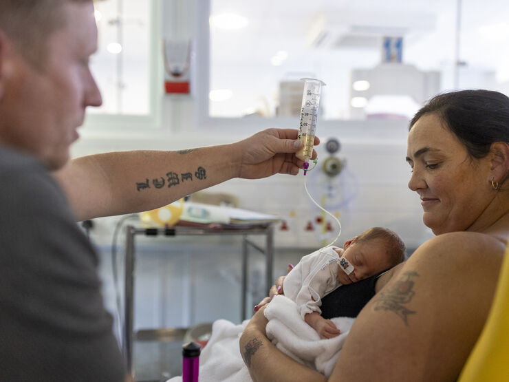 Mum holding baby on her chest while dad holds feeding tube in the air above baby