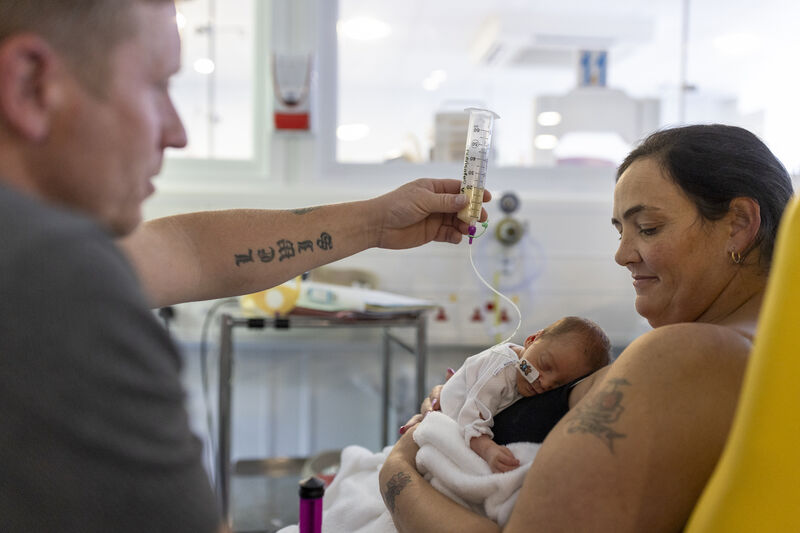 Mum holding baby on her chest while dad holds feeding tube in the air above baby