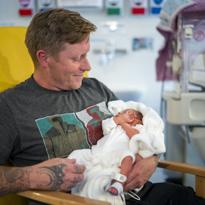 Dad sat in hospital chair holding his baby in his arms looking down and smiling