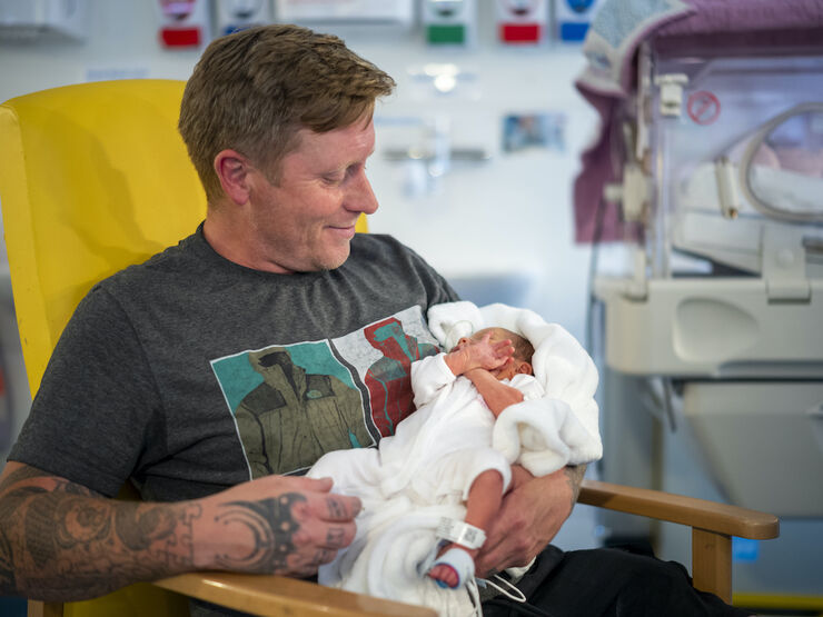 Dad sat in hospital chair holding his baby in his arms looking down and smiling