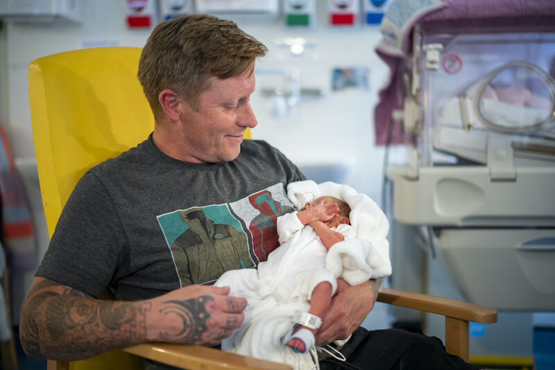 Dad sat in hospital chair holding his baby in his arms looking down and smiling