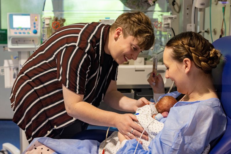 Woman holding baby on her chest in a hospital setting while her partner gently touches the baby's side