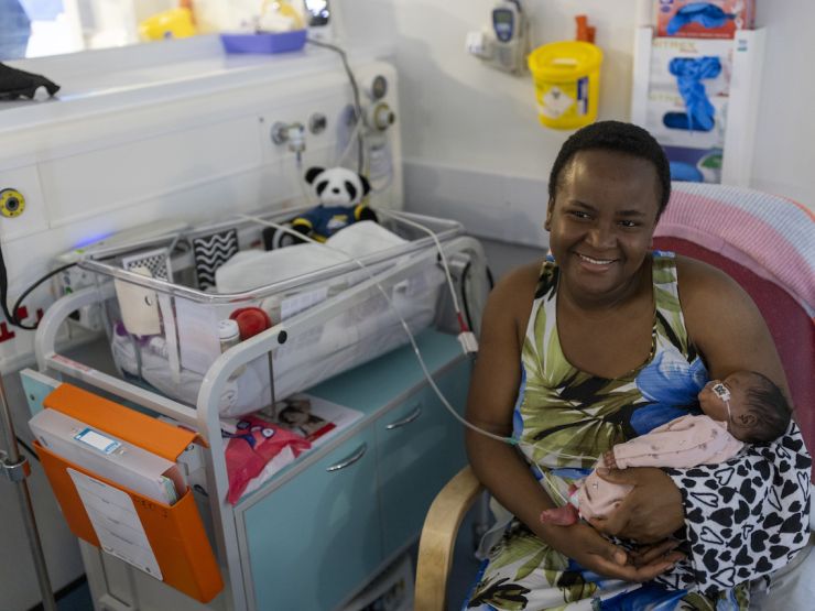 Mum smiling while holding her baby sat next to an incubator