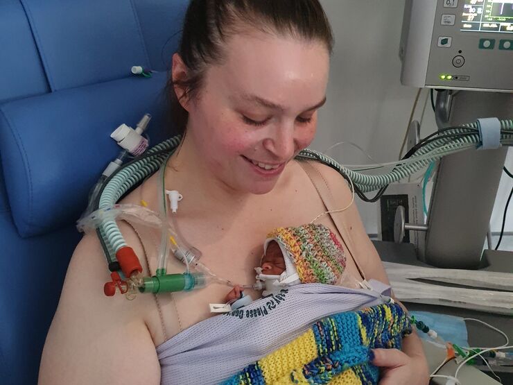 A parent in a hospital chair smiles looking down at their premature baby, who is directly against the skin on their chest, wrapped in a blanket. The baby has some medical tubes and wires attached to them.