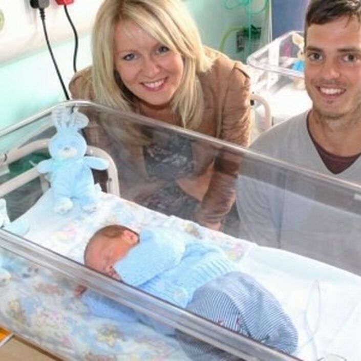 Dad and Esther Mcvey MP standing behind a cot with a premature baby smiling at the camera
