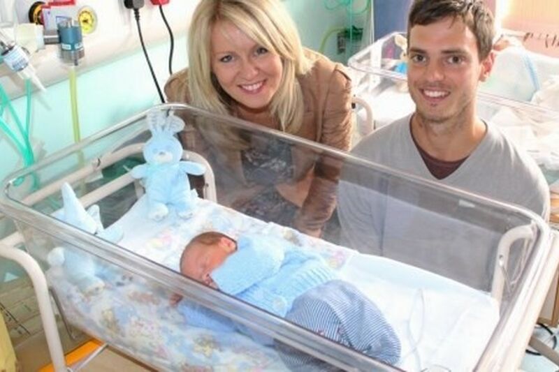 Dad and Esther Mcvey MP standing behind a cot with a premature baby smiling at the camera