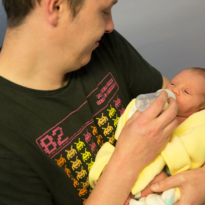Father sitting in a hospital chair, bottle feeding his baby who is wrapped in a yellow blanket