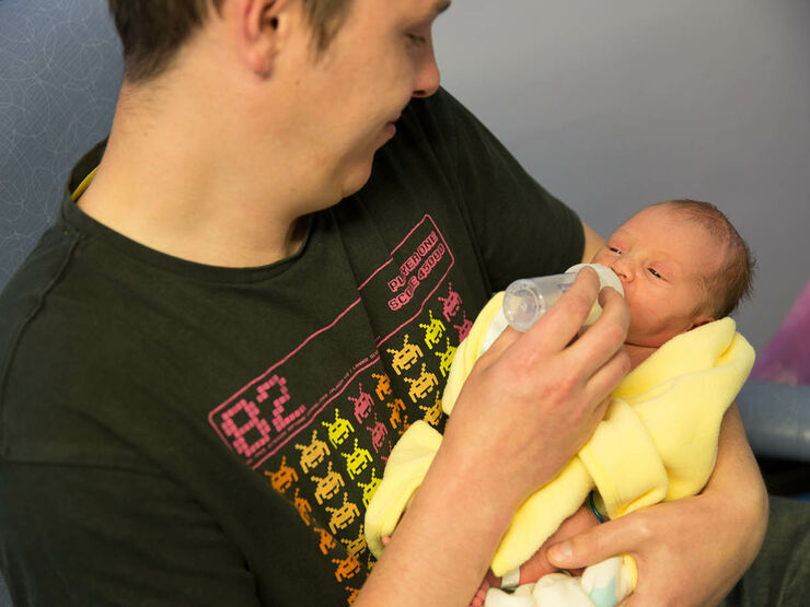 Father sitting in a hospital chair, bottle feeding his baby who is wrapped in a yellow blanket