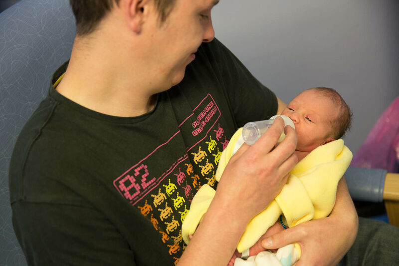 Father sitting in a hospital chair, bottle feeding his baby who is wrapped in a yellow blanket