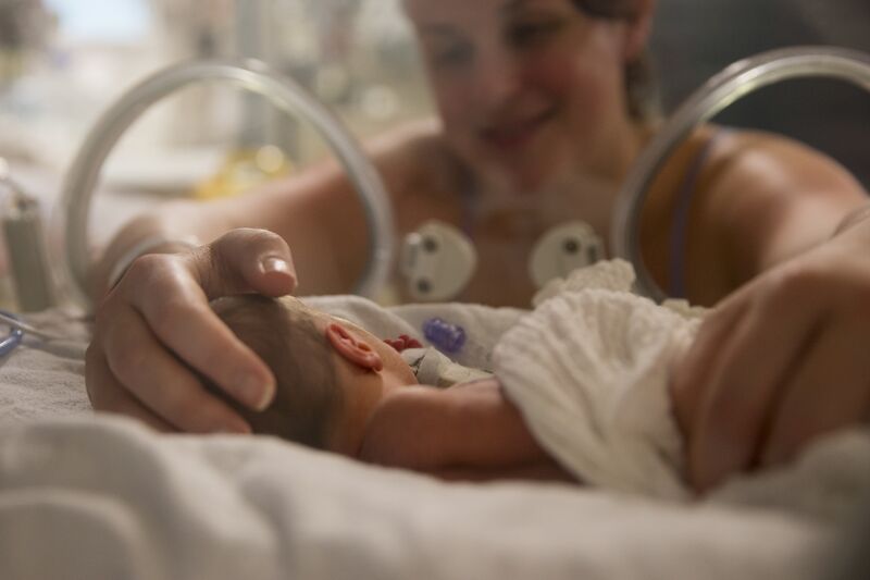 Baby in foreground in an incubator while mum in background has her hands through the incubator holding her baby's head