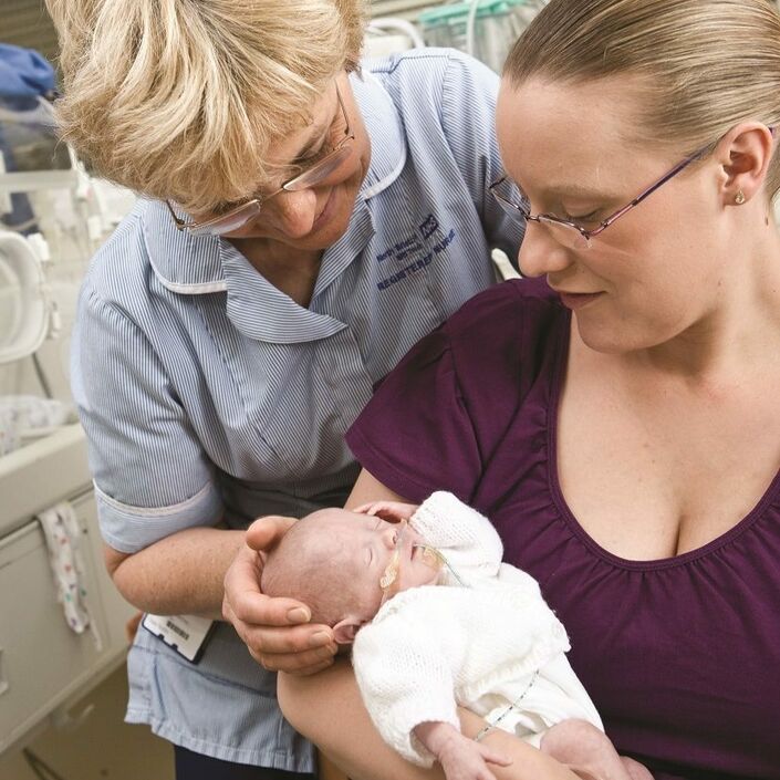 Mum holding baby in her arms with a nurse standing behind her with her hand supporting the baby's head