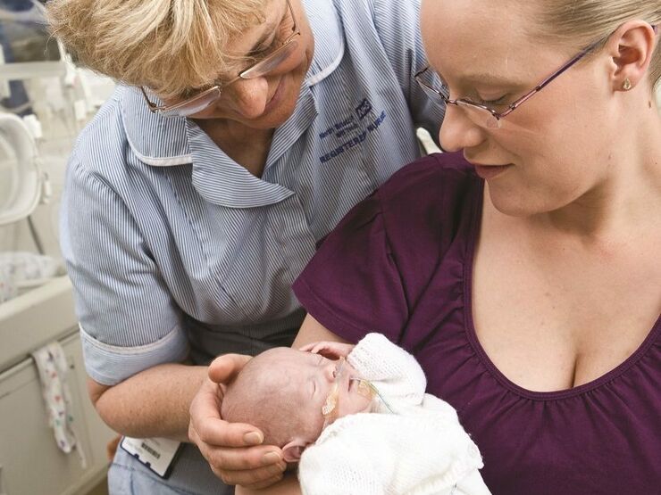Mum holding baby in her arms with a nurse standing behind her with her hand supporting the baby's head