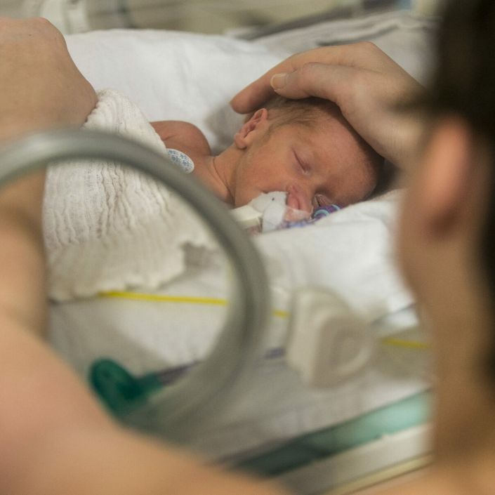Premature baby in incubator with dad putting his hands in and gently touching baby's head