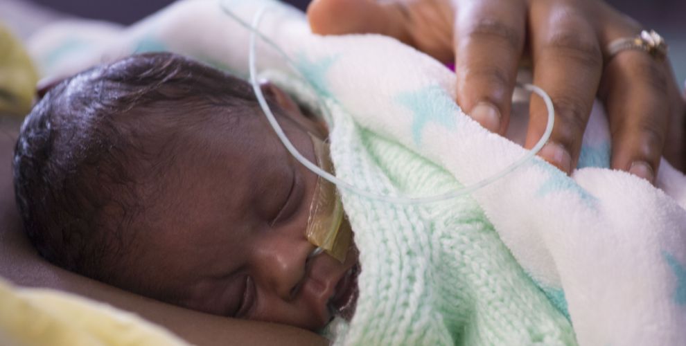 Premature baby wrapped in blanket sleeping on parent's chest with parent's hand on baby's back