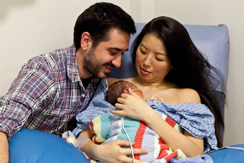 Mum holding baby on a hospital chair with dad alongside looking at baby. Baby is snuggled on mum's chest covered by a blanket.