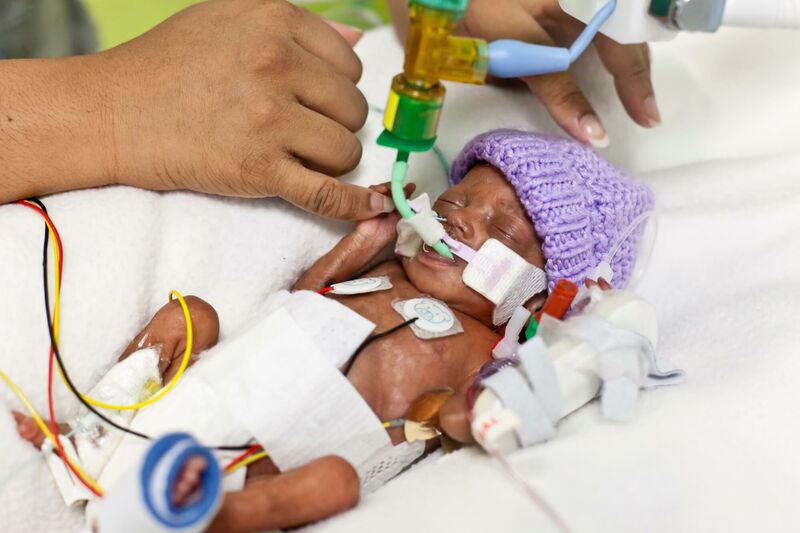 Premature baby in hospital wearing wool hat and attached to tubes holding parent's finger