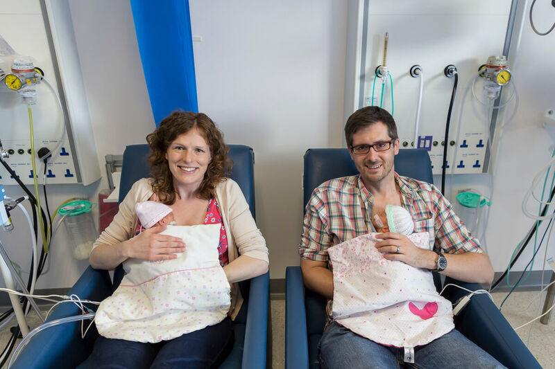 Mum and dad sat in hospital chairs smiling while holding twin babies