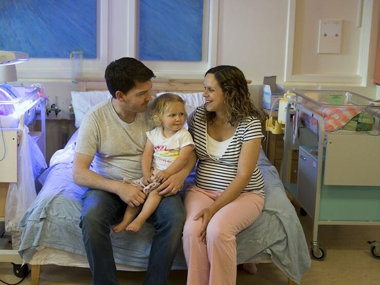 Parents sitting on a bed in hospital with cot beside them and with their toddler on dad's lap