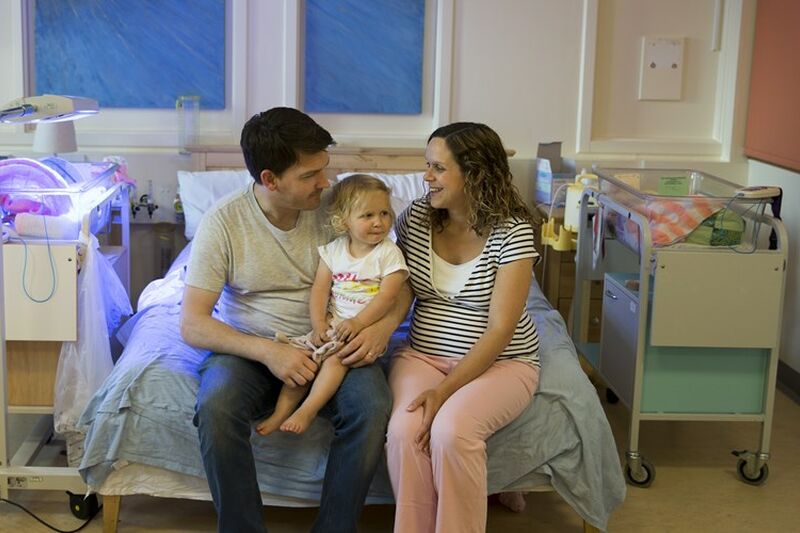 Parents sitting on a bed in hospital with cot beside them and with their toddler on dad's lap