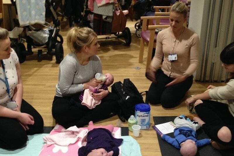 Group of mothers sat on the floor with two babies lying down while one baby being bottle fed
